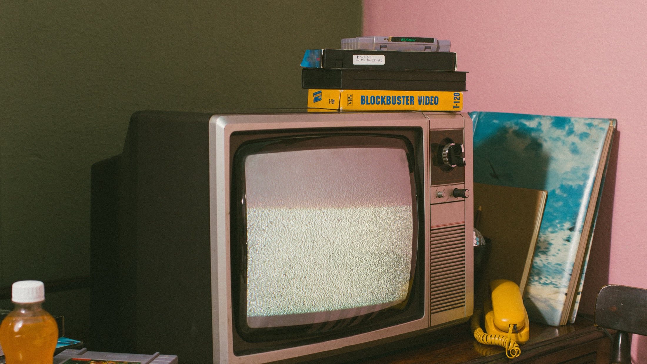 A cathode-ray television showing static, stacked VHS tapes and a yellow telephone in a lamplit room